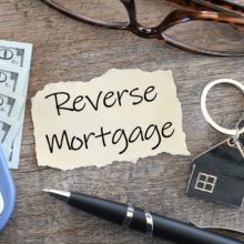 Paper with the words reverse mortgage on it along with a calculator, keys, pen, and glasses on a wooden table.